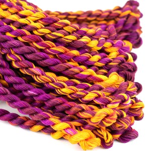 May include: A close-up of a pile of embroidery floss in shades of purple, orange, and yellow. The floss is twisted together in a rope-like shape.