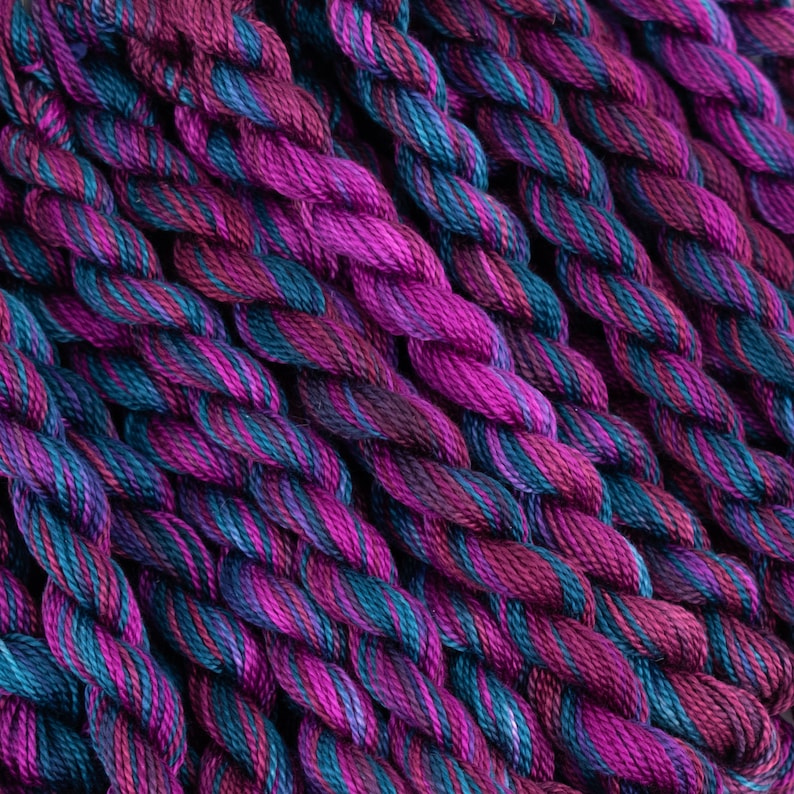 K&ouml;nnte beinhalten: Nahaufnahme eines B&uuml;ndels Stickgarn in Violett- und Blaut&ouml;nen. Das Garn ist miteinander verdreht und erzeugt ein strukturiertes und farbenfrohes Muster.