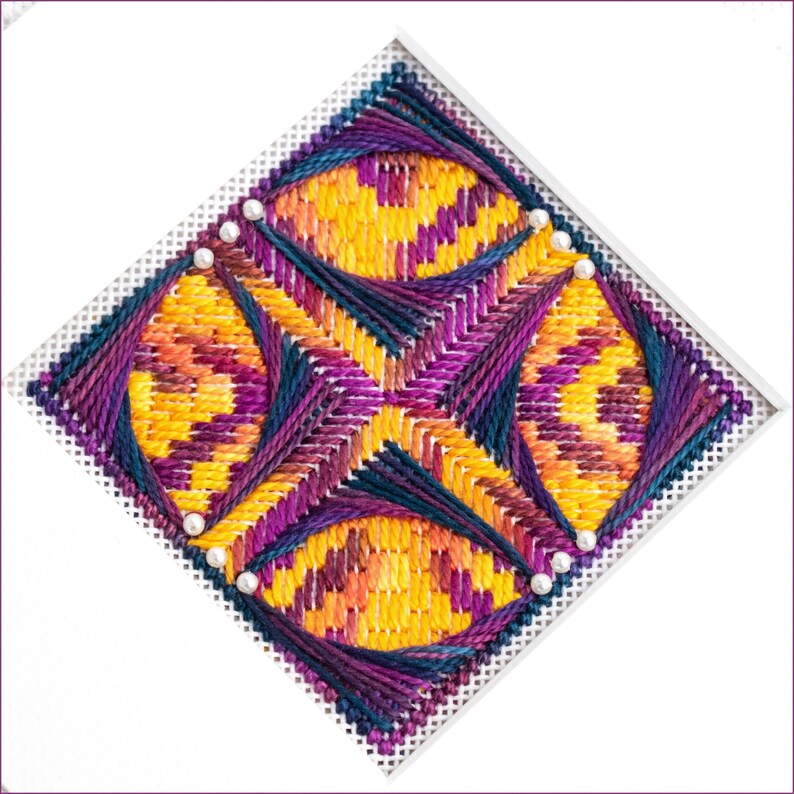 May include: A colorful cross-stitch design with a geometric pattern of overlapping circles in shades of yellow, orange, purple, and blue. The design is stitched on white Aida cloth and accented with white beads.
