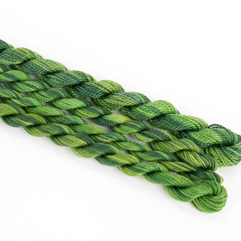 May include: Three skeins of green embroidery floss, each with a different shade of green. The floss is twisted together and arranged in a diagonal line.