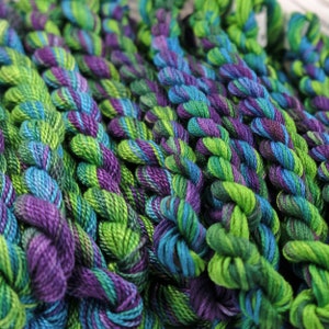 May include: Close-up of a collection of embroidery floss spools in shades of green, purple, and blue. The floss is twisted and textured, creating a visually appealing pattern.