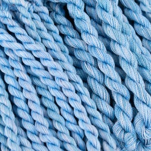 May include: A collection of light blue twisted cotton embroidery threads arranged in parallel vertical strands. The fibres exhibit a variegated colour pattern with subtle shifts between pale sky blue and white, creating a textured appearance.