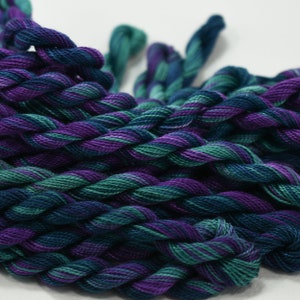 May include: Close-up of several skeins of embroidery floss in shades of teal, purple, and blue. The floss is twisted into a rope-like pattern, with the colors blending together. The background is plain white.