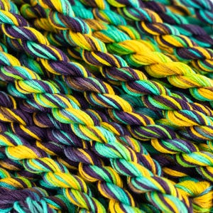 May include: Close-up of a bundle of embroidery floss in shades of green, yellow, and purple.