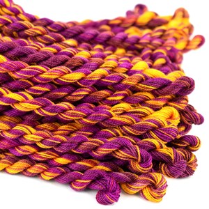 May include: A close-up of a pile of embroidery floss in shades of purple and yellow. The floss is twisted together in a rope-like shape.
