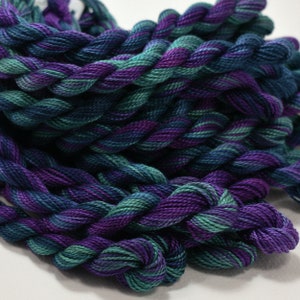May include: Close-up of a bundle of embroidery floss in shades of teal, purple, and navy blue. The threads are twisted together, creating a textured appearance. The colors blend together, creating a gradient effect.