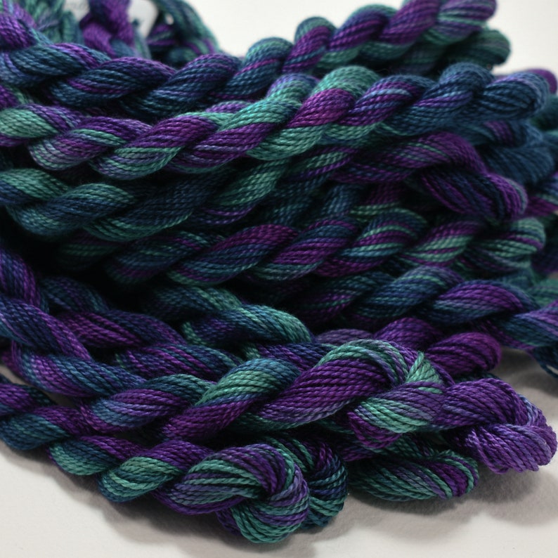 May include: Close-up of a bundle of embroidery floss in shades of teal, purple, and navy blue. The threads are twisted together, creating a textured appearance. The colors blend together, creating a gradient effect.