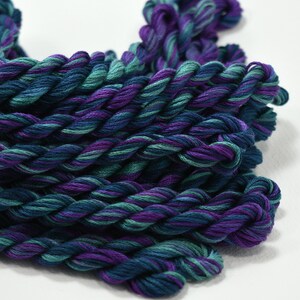 May include: Close-up of several skeins of embroidery floss in shades of teal, purple, and blue. The floss is tightly twisted, creating a textured appearance. The colors are blended, giving a gradient effect. Ideal for embroidery, cross-stitch, or other needlecraft projects.