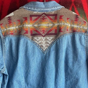 Pode incluir: Camisa jeans azul clara com um jugo decorativo estampado. O jugo apresenta um design geométrico em tons de castanho, laranja, vermelho e creme. A camisa tem mangas compridas e punhos abotoados.