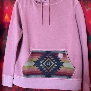 Op de afbeelding: Zachtroze hoodie met een kangoeroezak met een geometrisch patroon geïnspireerd op het zuidwesten in groen, rood en marineblauw. De hoodie heeft een capuchon met trekkoord en geribde manchetten en zoom. Een klein logo is zichtbaar.