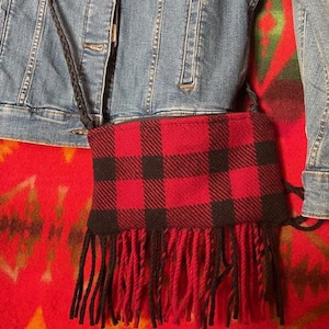 Op de afbeelding: Een klein rood en zwart geruit crossbody tasje met een franje rand. De tas heeft een zwarte band en is gemaakt van een zachte, geweven stof.