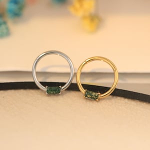 Puede incluir: Dos aros nasales circulares, uno plateado y otro dorado, cada uno con una gema verde rectangular. Los aros se exhiben sobre una superficie neutra, mostrando su diseño y contraste de color. Ideales para joyería corporal.