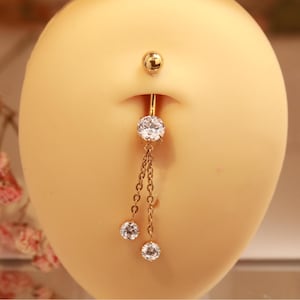 Puede incluir: Un piercing para el ombligo de color dorado con tres gemas redondas y brillantes. La bola superior y las dos gemas colgantes están conectadas por una delicada cadena. La joya se exhibe sobre una superficie clara.