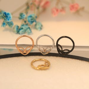 May include: Four nose rings in different metallic finishes: rose gold, silver, gold, and black. Each ring features an alien head design. The rings are displayed on a light surface with blurred floral elements in the background.
