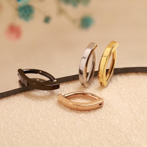 May include: Four belly button rings in different metallic finishes: black, silver, gold, and rose gold. Each ring has a curved, oval shape with a flat bar across the top. The rings are displayed on a light-colored surface.