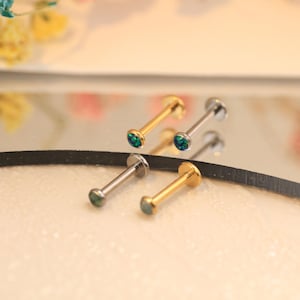 Peut inclure: Quatre piercings pour les lèvres. Deux sont de couleur or et deux sont de couleur argent. Chaque piercing a une petite pierre précieuse ronde verte et bleue. Les piercings sont présentés sur une surface réfléchissante.