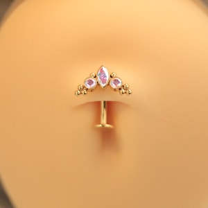 May include: A gold-coloured belly button ring with a decorative top. The top features a central marquise-shaped iridescent gem flanked by two round gems and small gold beads. The ring is designed for body piercing.