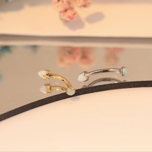 Puede incluir: Dos piercings curvos para cejas. Uno es dorado, el otro plateado. Cada anillo presenta una pequeña piedra de ópalo blanco y redonda en cada extremo. Las joyas se exhiben sobre una superficie reflectante.