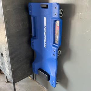 May include: A blue automotive creeper with black wheels and the text "PITTSBURGH AUTOMOTIVE" printed on the side. The creeper is mounted to a wall.