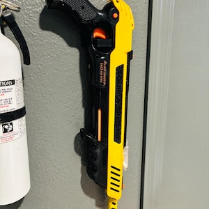 May include: A black and yellow stun gun is mounted on a gray wall. The stun gun has a black handle and a yellow body with the text "DO NOT SHOOT IN FACE OR EYES!". An orange tip is visible. A fire extinguisher is to the left.