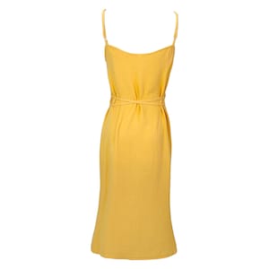 May include: A mustard yellow midi dress featuring delicate spaghetti straps and a matching waist tie. The dress is crafted from a textured material and has a simple, elegant design. The dress is a solid colour, suitable for warmer weather.