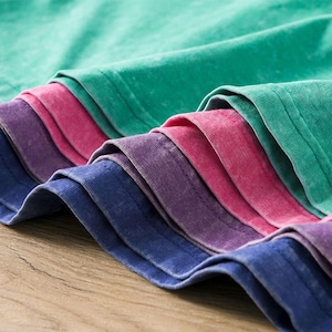 Op de afbeelding: Close-up van een stapel gevouwen t-shirts in de kleuren groen, roze, paars en blauw. De naden en de textuur van de stof zijn zichtbaar. De kleuren zijn levendig en de stof voelt zacht aan.
