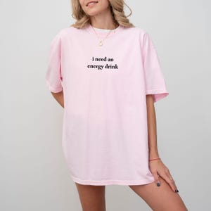 May include: A light pink t-shirt with the text "i need an energy drink" in black. The shirt has short sleeves and a relaxed fit. The model is wearing a gold necklace and a red bracelet.
