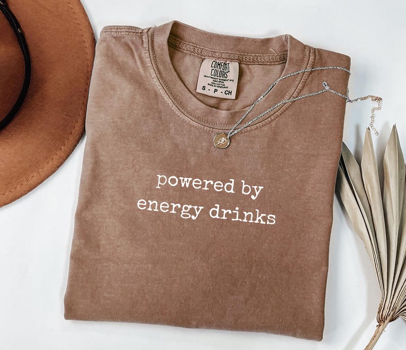 K&ouml;nnte beinhalten: Ein braunes T-Shirt mit dem wei&szlig;en Text "powered by energy drinks". Eine silberne Halskette mit einem kleinen Anh&auml;nger liegt auf dem Shirt. Ein brauner Hut und ein getrocknetes Palmblatt befinden sich im Hintergrund.