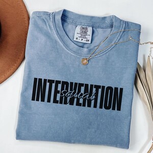 May include: A light blue Comfort Colors t-shirt with the words "INTERVENTION squad" printed in black. A gold necklace with a pendant is draped across the shirt. A brown hat and dried plant are in the background.