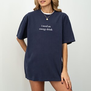 May include: Navy blue short-sleeve t-shirt with the text "i need an energy drink" in white. The shirt has a relaxed fit and a white undershirt is visible at the neckline. A gold necklace is also visible.