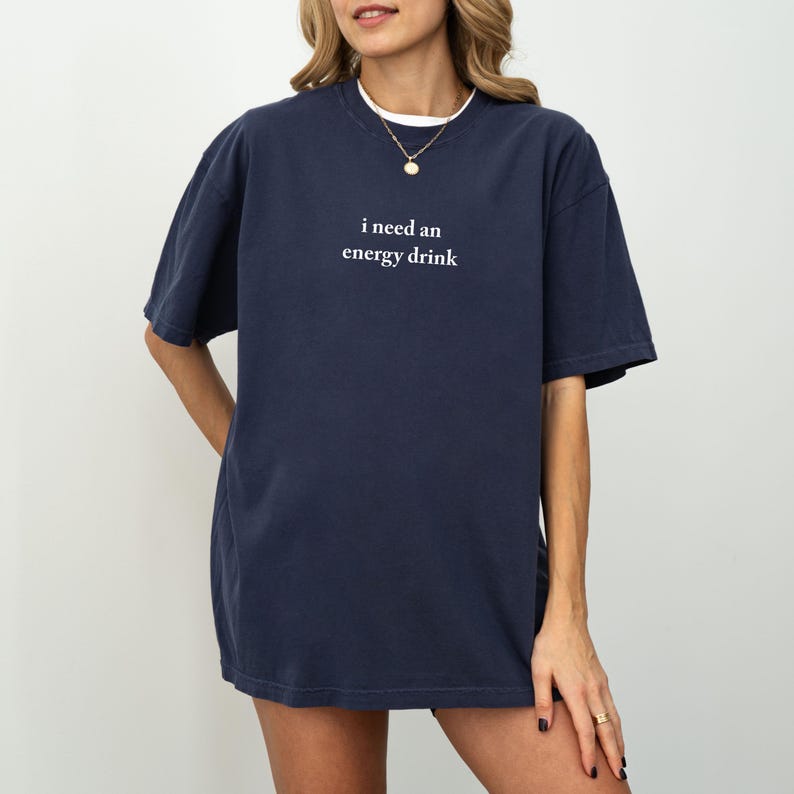 May include: Navy blue short-sleeve t-shirt with the text "i need an energy drink" in white. The shirt has a relaxed fit and a white undershirt is visible at the neckline. A gold necklace is also visible.