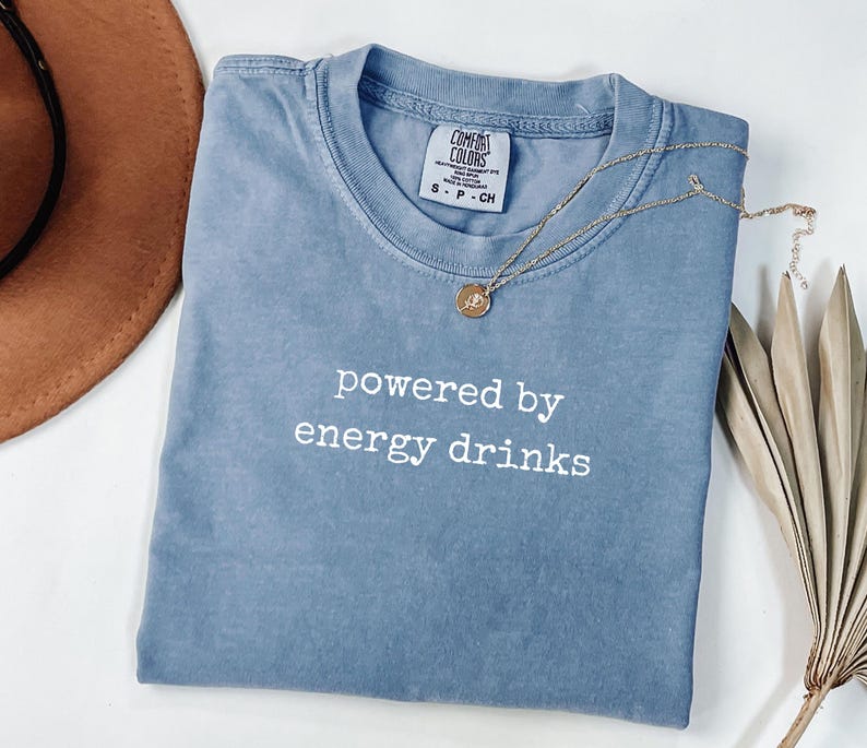 K&ouml;nnte beinhalten: Ein blaues T-Shirt mit kurzen &Auml;rmeln und dem wei&szlig;en Text "powered by energy drinks". Eine goldene Halskette mit einem kleinen Anh&auml;nger befindet sich auf dem Shirt. Das Shirt ist auf einer wei&szlig;en Oberfl&auml;che gefaltet.