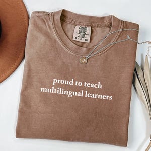May include: A brown Comfort Colors t-shirt with the text "proud to teach multilingual learners" in white. A silver necklace with a pendant is draped across the shirt. A brown hat and dried palm leaf are in the background.