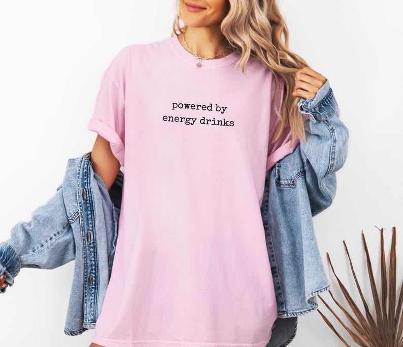 K&ouml;nnte beinhalten: Hellrosa T-Shirt mit dem schwarzen Text "powered by energy drinks". Das Shirt wird mit einer blauen Jeansjacke getragen.