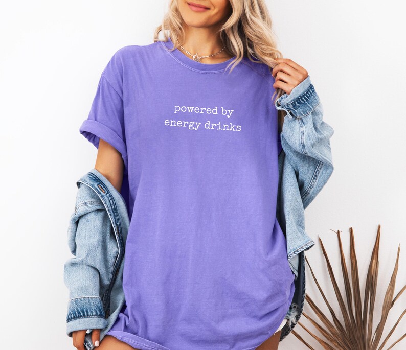 K&ouml;nnte beinhalten: Ein lila T-Shirt mit dem wei&szlig;en Text "powered by energy drinks". Das Shirt hat hochgekrempelte &Auml;rmel und wird mit einer hellblauen Jeansjacke getragen. Der Stil ist l&auml;ssig und entspannt.