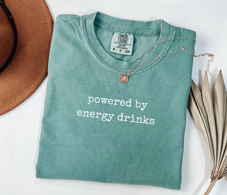 K&ouml;nnte beinhalten: Ein t&uuml;rkisfarbenes Comfort Colors T-Shirt mit dem wei&szlig;en Schriftzug "powered by energy drinks". Eine silberne Halskette mit einem runden Anh&auml;nger liegt auf dem Shirt. Ein brauner Hut und eine getrocknete Pflanze befinden sich im Hintergrund.