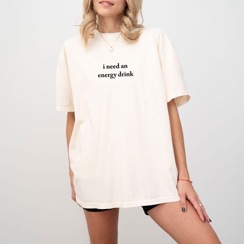 May include: Cream-colored t-shirt with the text "i need an energy drink" in black, centered on the front. The shirt has short sleeves and a relaxed fit. The model is wearing black shorts.