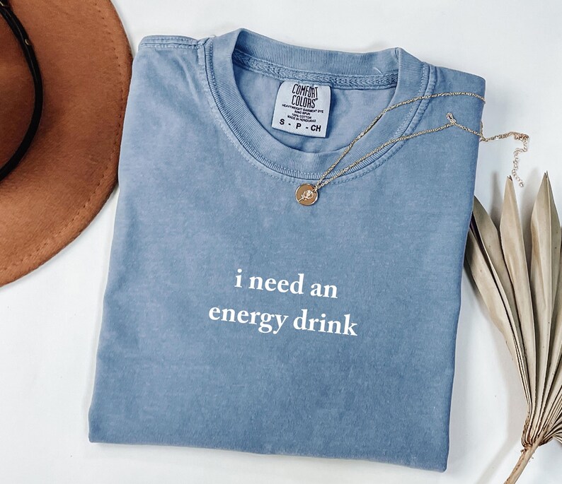 May include: A blue Comfort Colors t-shirt with the text "i need an energy drink" in white. A gold necklace with a pendant is draped across the shirt. A brown hat and dried plant are in the background.