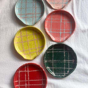 Op de afbeelding: Een verzameling van zes keramische lepelsteunen in verschillende kleuren: lichtblauw, roze, rood, geel en groen. Elke steun heeft een ruitpatroon in een contrasterende kleur en gespikkelde details. De lepelsteunen zijn rond met een gebogen handgreep.