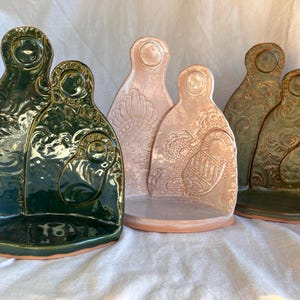 May include: A collection of ceramic sculptures in shades of green and peach. The sculptures have a stylized human form with embossed patterns. The pieces are arranged on a white surface, showcasing their unique shapes and textures.