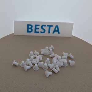 Könnte beinhalten: Ein Haufen weißer Möbelstopfen aus Kunststoff, auch bekannt als Möbelbefestigungen, mit dem Wort "BESTA" auf einer weißen Karte im Hintergrund.