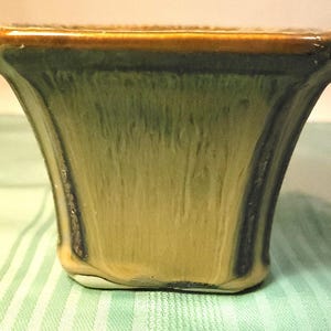 Könnte beinhalten: Eine quadratische Keramikvase mit ausgestellter Form. Die Vase hat eine grüne und beige Glasur mit einem braunen Rand. Die Glasur hat einen Tropfeffekt, der ein strukturiertes Aussehen erzeugt. Die Vase steht auf einer grün-weiß gestreiften Oberfläche.