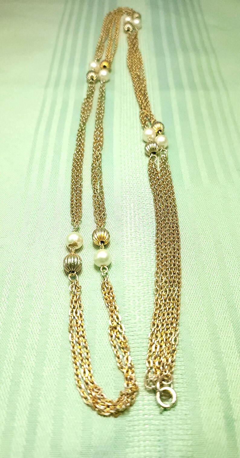 Puede incluir: Collar dorado con m&uacute;ltiples hilos, con peque&ntilde;as perlas y cuentas decorativas. El collar se muestra sobre un fondo verde claro texturizado, mostrando su delicado dise&ntilde;o y los intrincados detalles de la cadena.