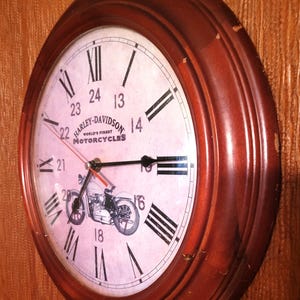 Könnte beinhalten: Runde Wanduhr aus Holz mit hellem Zifferblatt, das das Harley-Davidson-Logo und eine Motorrad-Illustration zeigt. Die Uhr hat schwarze römische Ziffern und Zeiger, mit einem roten Sekundenzeiger. Der Rahmen ist dunkelbraun. Durchmesser ca. 30 cm.