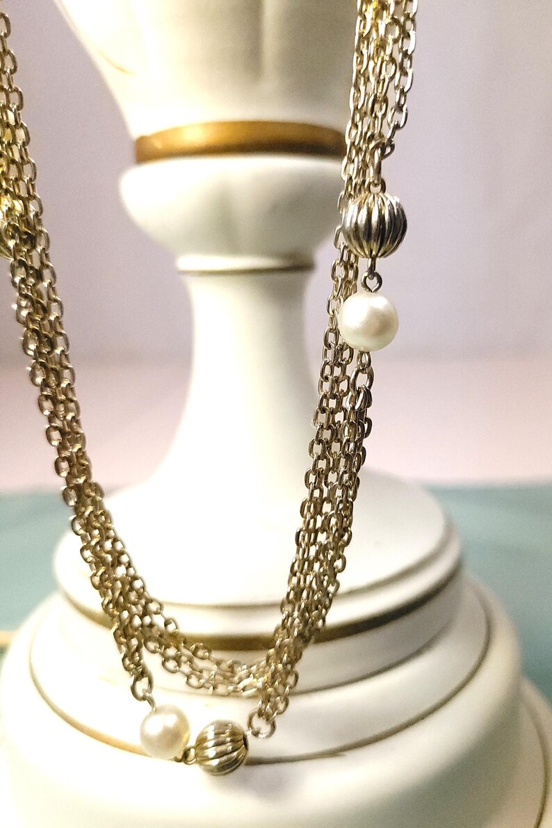 Puede incluir: Un collar dorado con m&uacute;ltiples hilos, con una combinaci&oacute;n de eslabones de cadena, cuentas de metal acanaladas y acentos de perlas blancas. El collar se muestra sobre un fondo blanco.