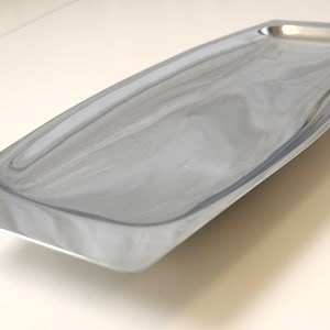 Könnte beinhalten: Ein rechteckiges, silberfarbenes Metalltablett mit einer polierten, reflektierenden Oberfläche. Das Tablett verfügt über eine flache, längliche Vertiefung mit leicht geschwungenen Seiten und flachen, zulaufenden Enden für ein glattes Design.