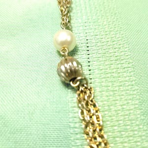 Puede incluir: Collar dorado con un dise&ntilde;o de cadena delicada. El collar presenta una perla blanca grande y redonda y una cuenta texturizada de color plateado. Un grupo de cadenas doradas cuelga de la parte inferior. El fondo es una tela verde claro.