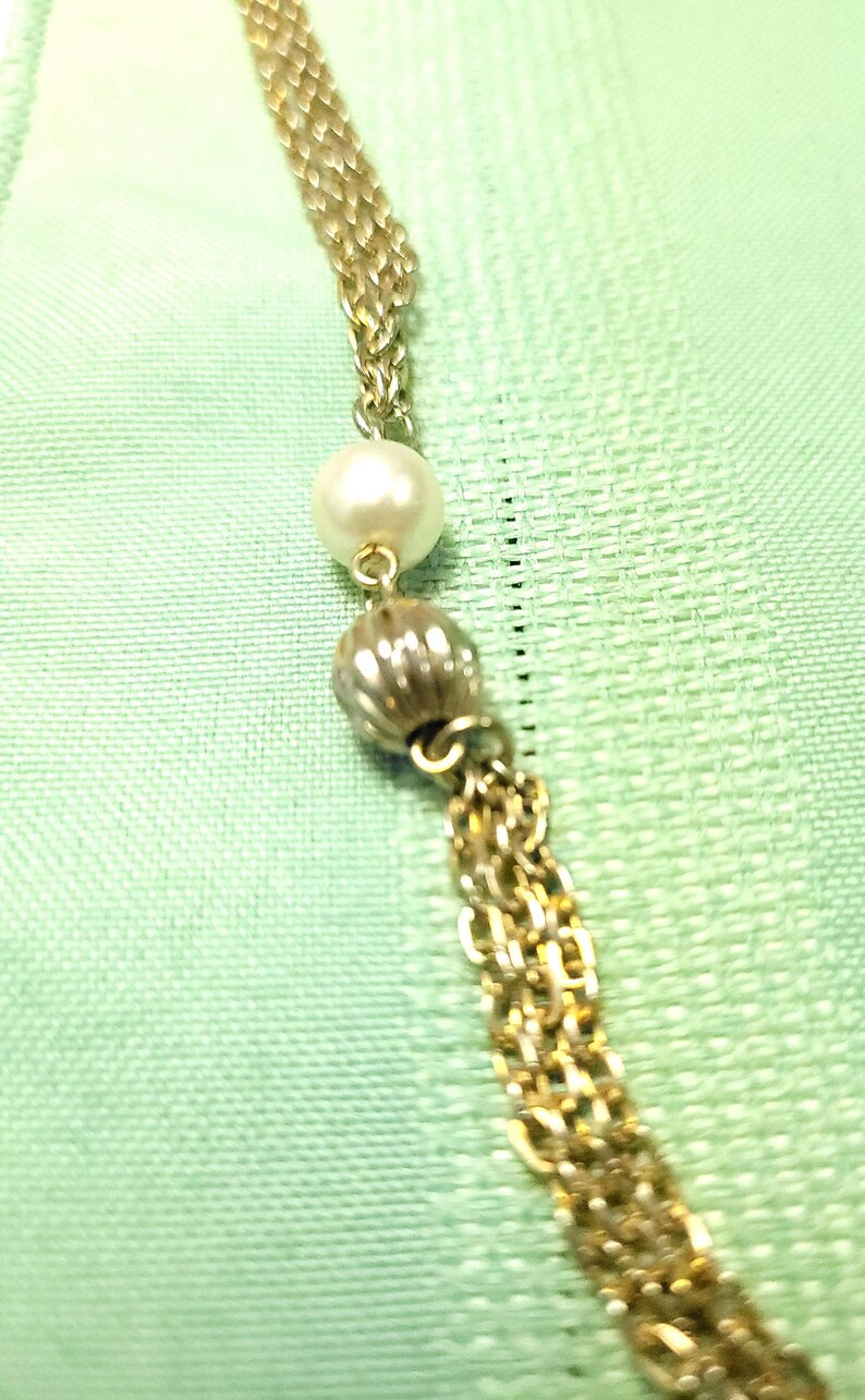 Puede incluir: Collar dorado con un dise&ntilde;o de cadena delicada. El collar presenta una perla blanca grande y redonda y una cuenta texturizada de color plateado. Un grupo de cadenas doradas cuelga de la parte inferior. El fondo es una tela verde claro.