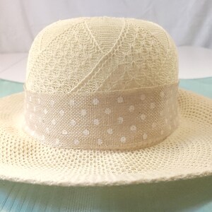 May include: A cream-colored sun hat with a wide brim. The crown features a delicate lace pattern, and a beige band with white polka dots encircles the base. The hat is designed for sun protection and style.