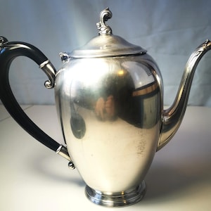 May include: A polished silver teapot with a black, curved handle and a decorative lid. The spout is elegantly curved. The teapot is designed with a classic shape, reflecting light across its surface. The handle is made of a dark, possibly ebonized, material.